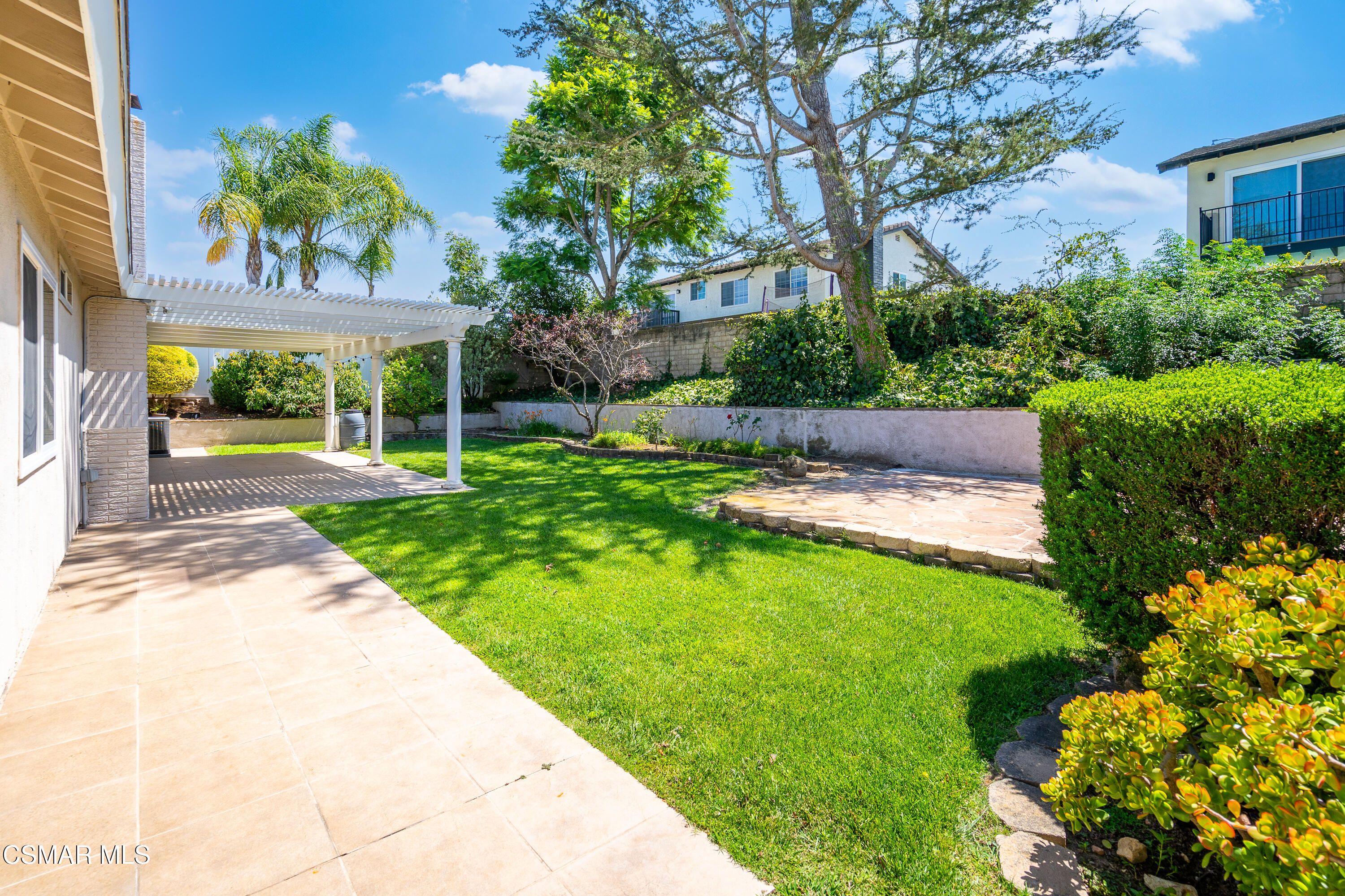 Backyard with a broad lawn, white pergola-covered patio, stone sitting area, and mature trees.