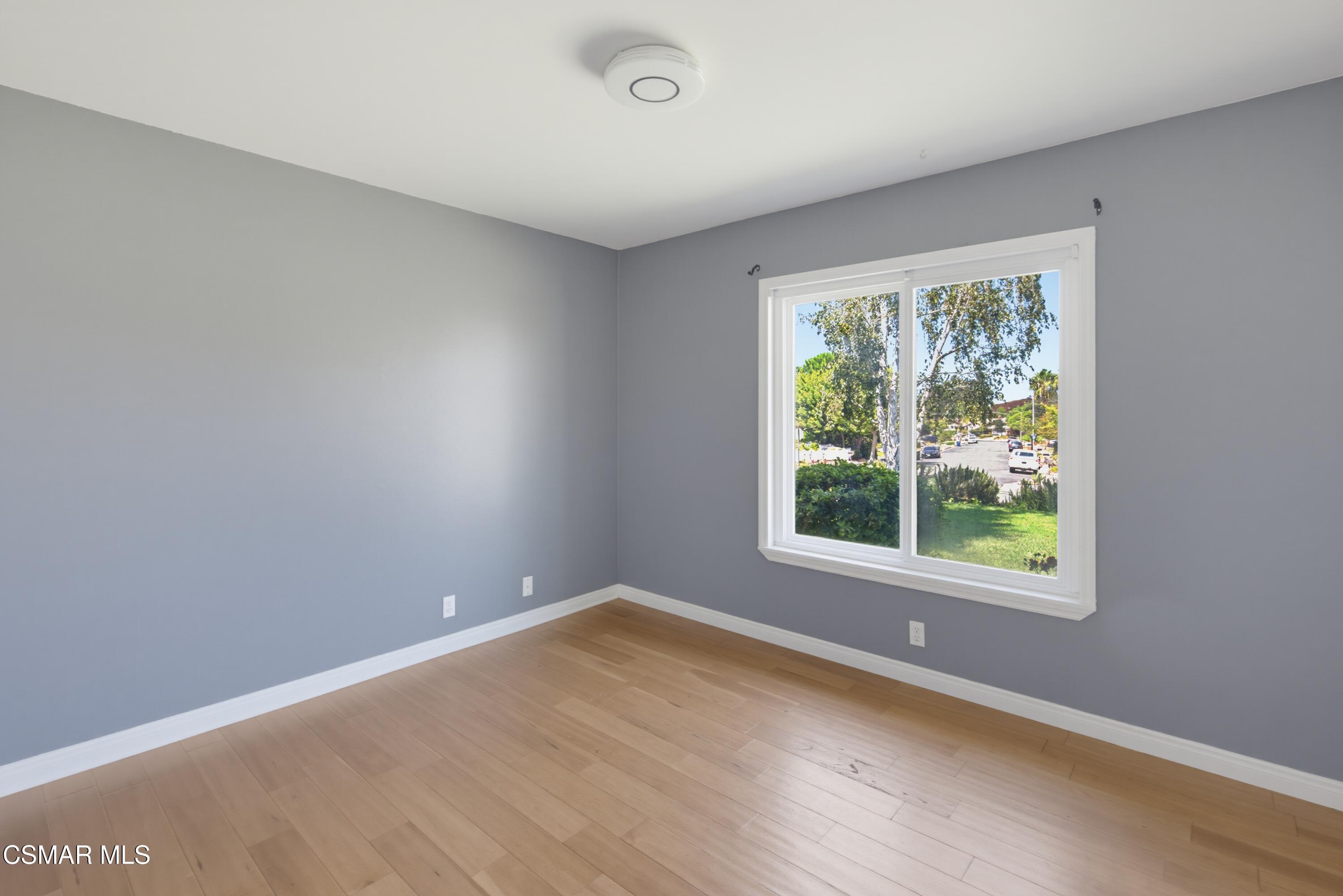 Empty room with gray walls, light wood floors, and a large window overlooking trees and a residential street.