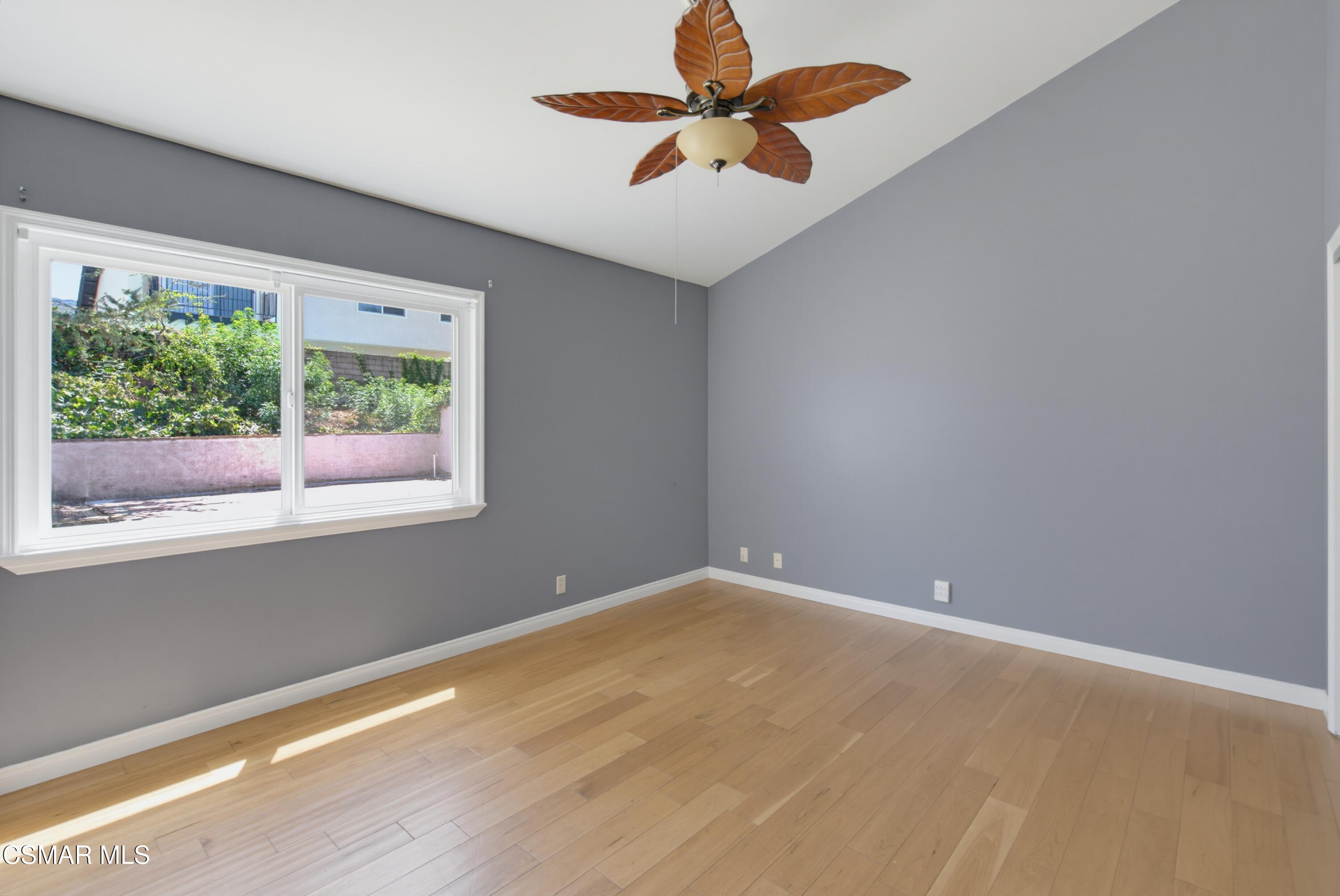 Empty gray-walled room with hardwood floors, a large window, and a leaf-blade ceiling fan.