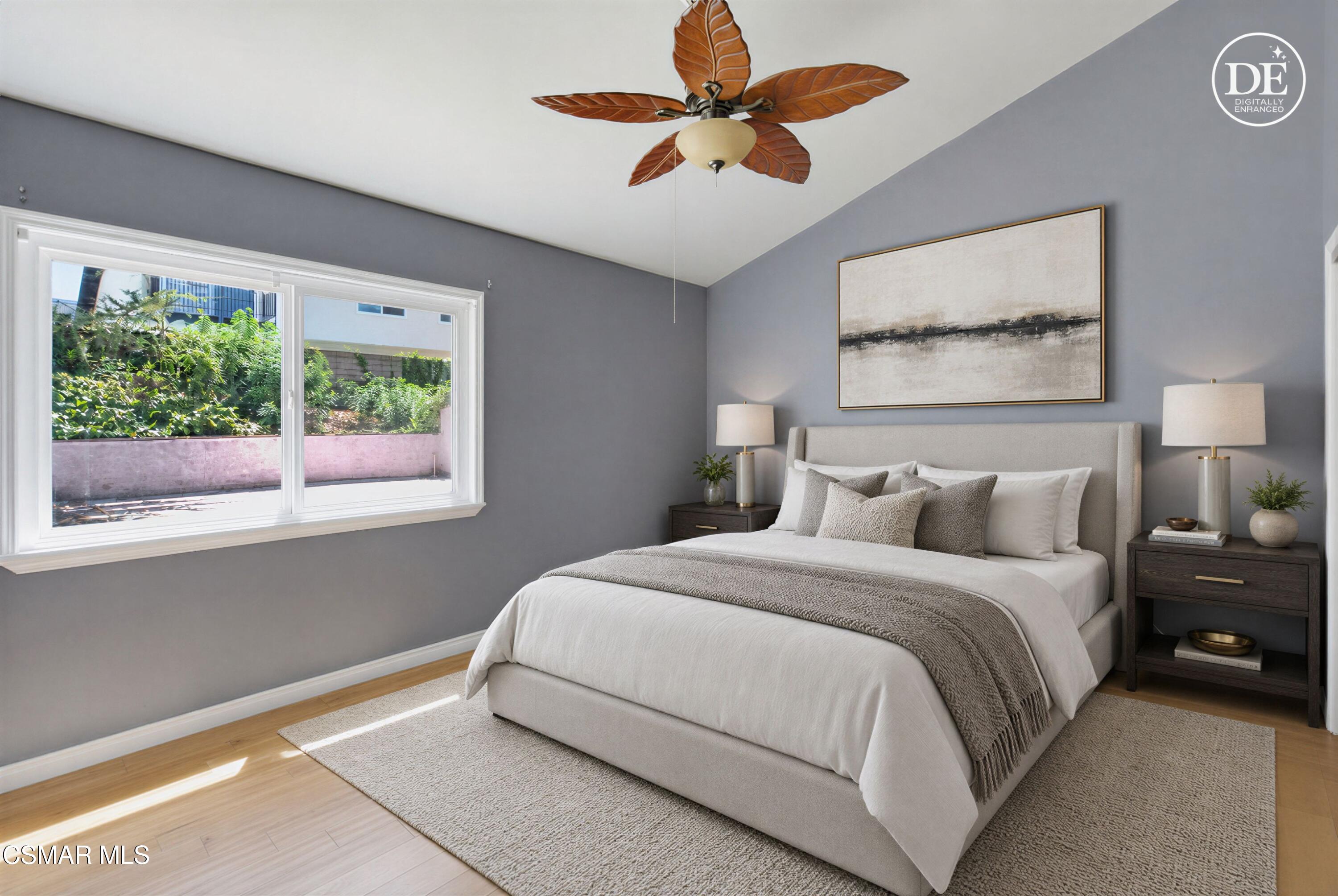 Staged bedroom with a neutral upholstered bed, gray walls, leaf-shaped ceiling fan, and sunlit window overlooking greenery.