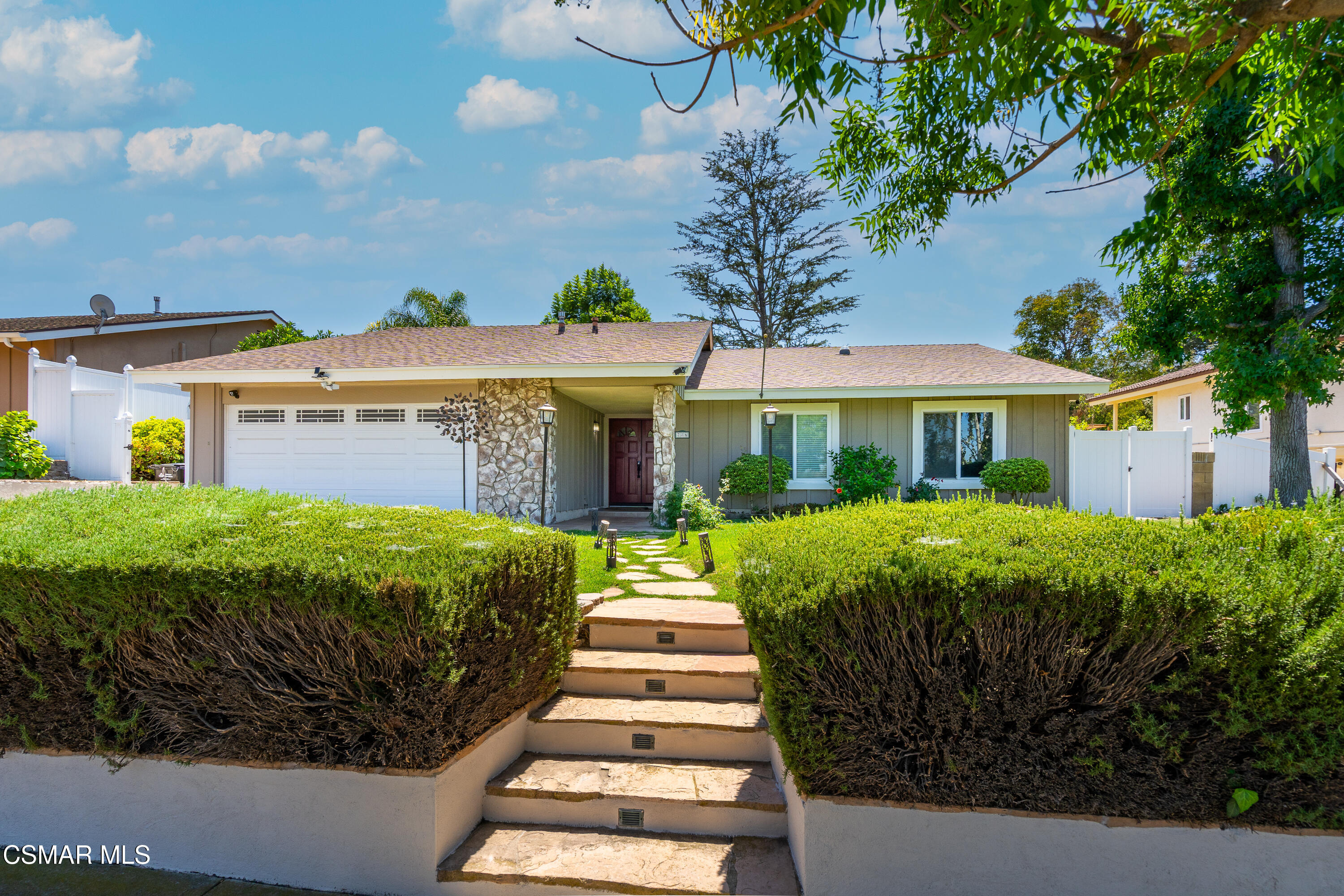 Single-story gray home with stone entry, two-car garage, and steps bordered by trimmed hedges.