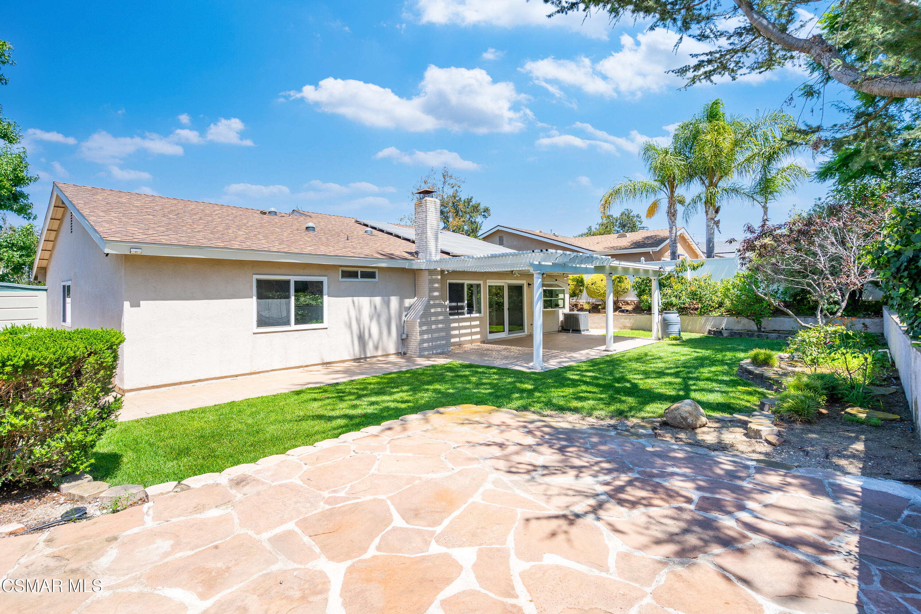 Home’s backyard with a covered patio, green lawn, landscaped garden, and broad flagstone terrace.