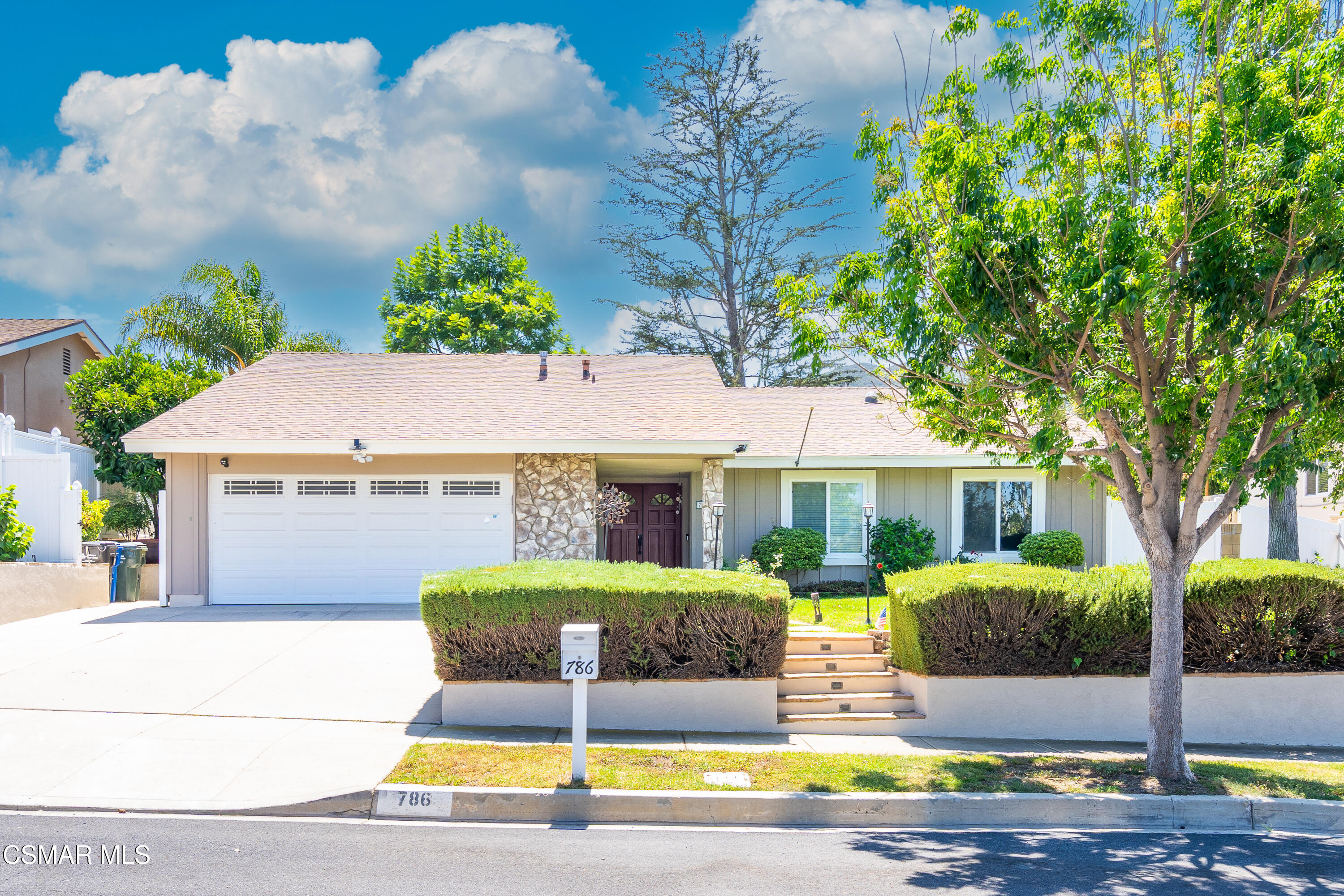 Single-story home with a wide garage, stone entry, clipped hedges, and a large shade tree.