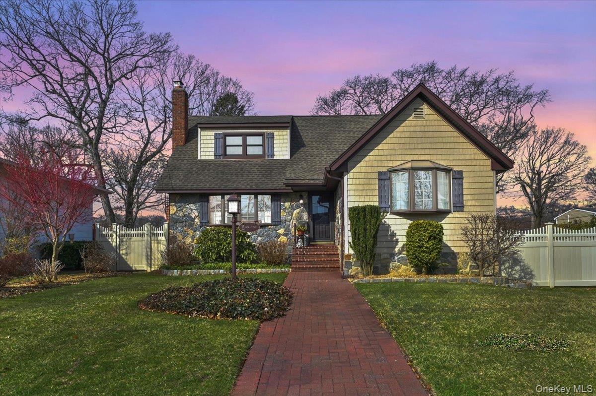 Yellow-shingled cottage with stone facade, brick walkway, and white fence at dusk.
