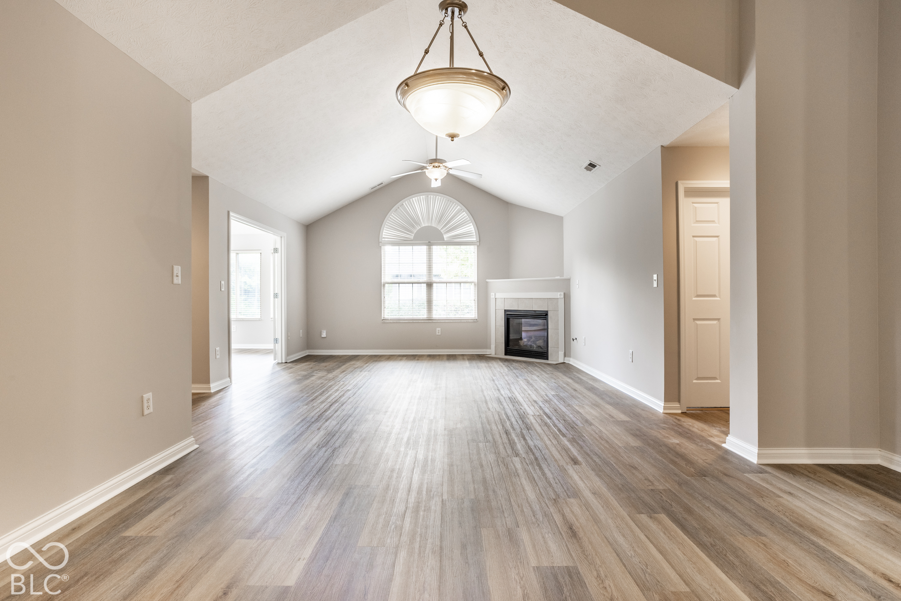 Empty living room with vaulted ceiling, arched window, fireplace, and wood-look flooring.