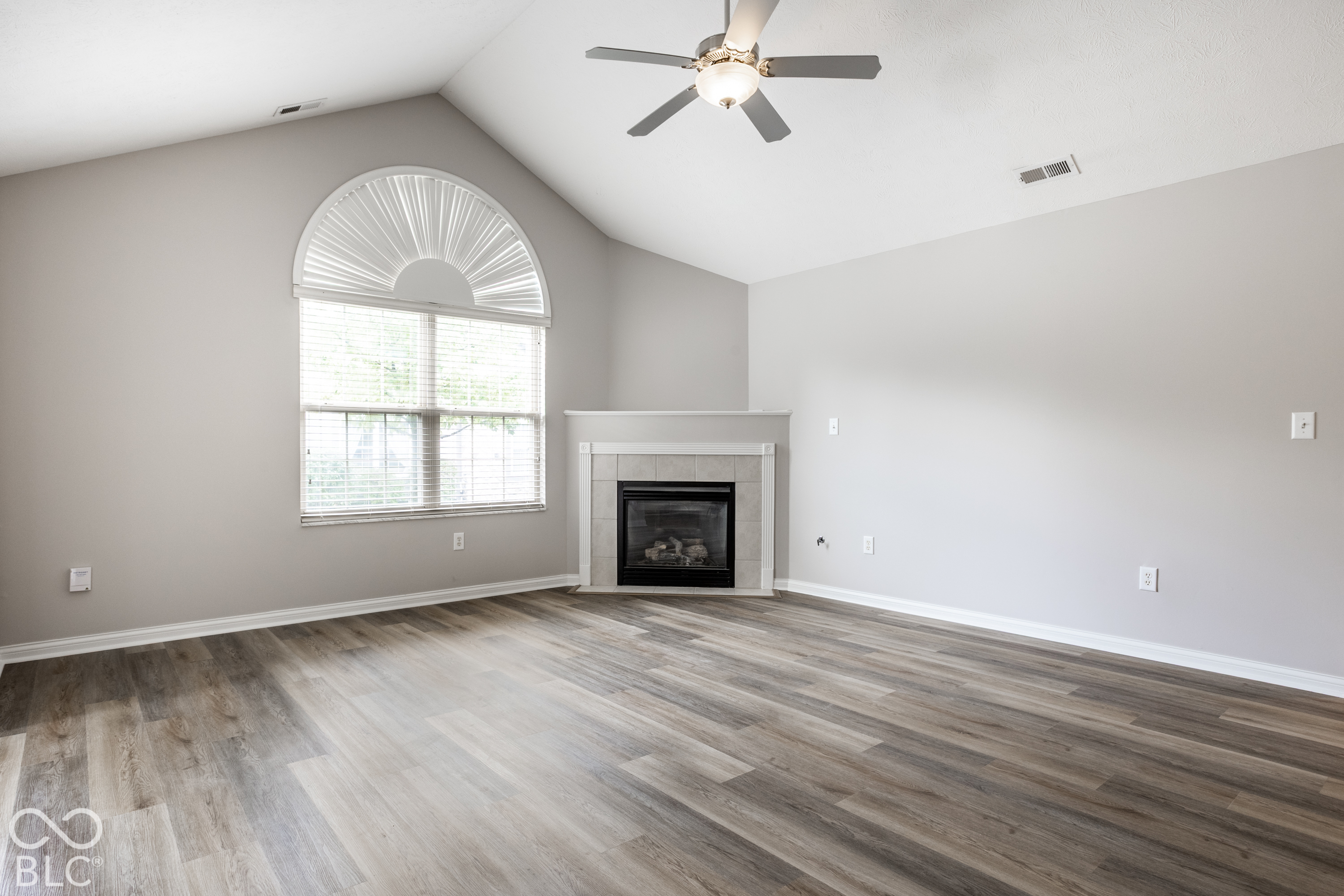 Empty living room with a tiled fireplace, arched window, and gray wood-look flooring.