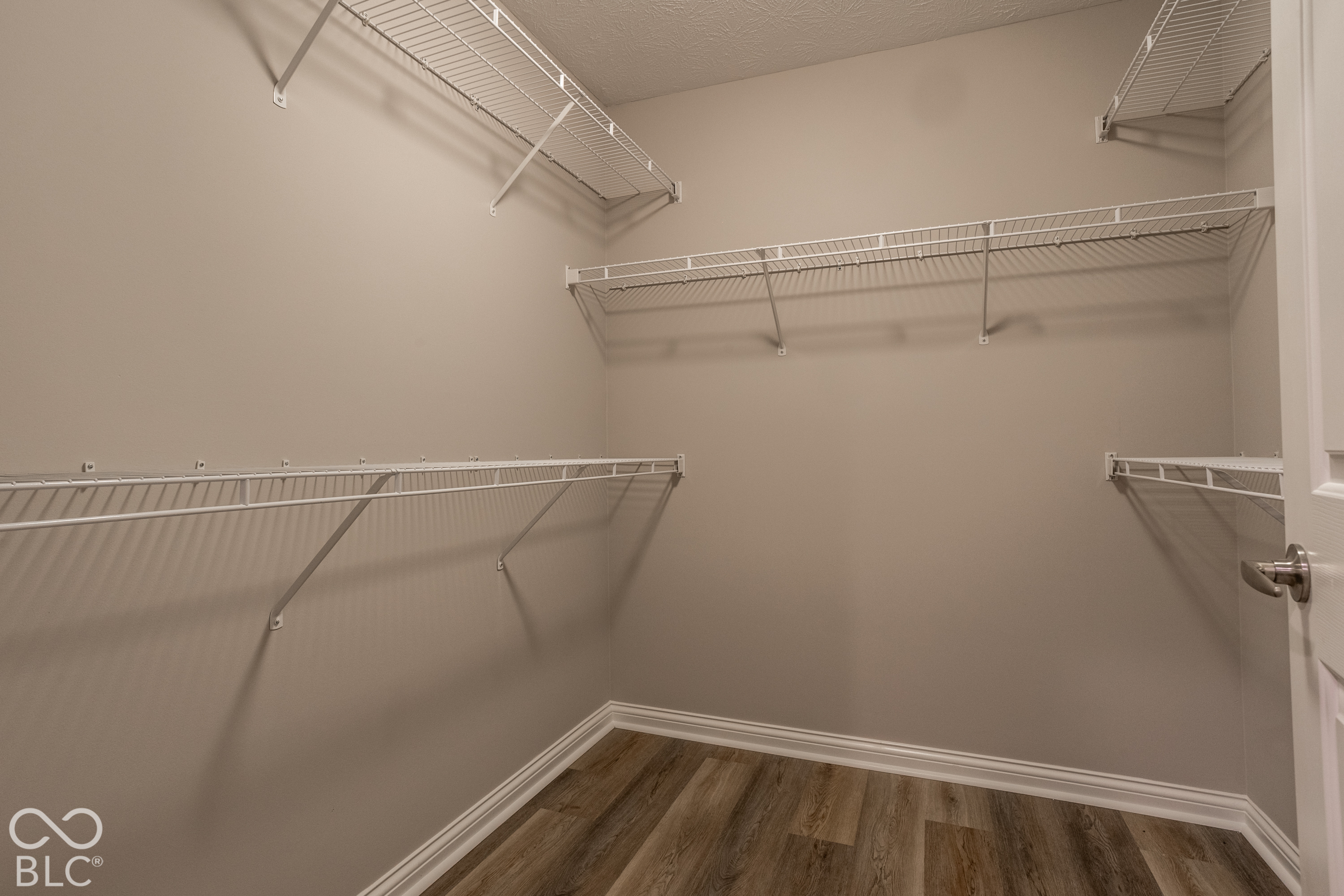 Empty walk-in closet with wraparound wire shelving and dark wood-look flooring.