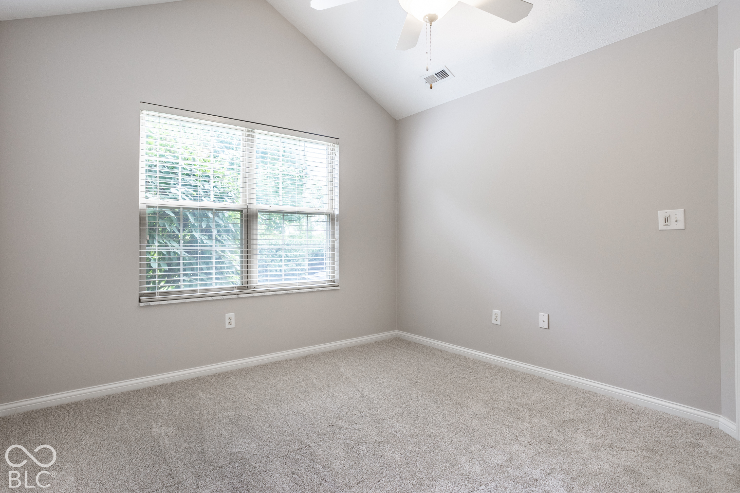 Empty bedroom with beige carpet, vaulted ceiling, ceiling fan, and a large window with blinds.