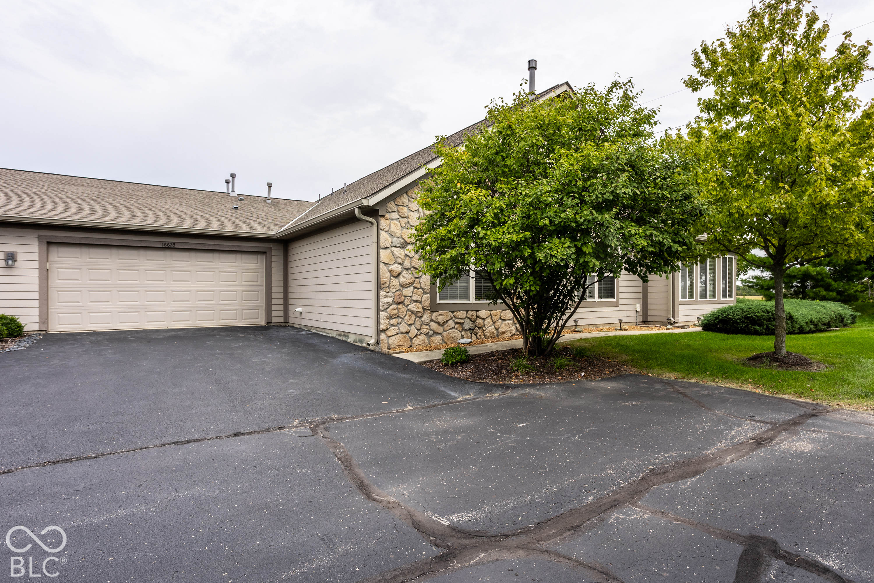Home exterior with a two-car garage, stone facade, broad driveway, and mature trees.