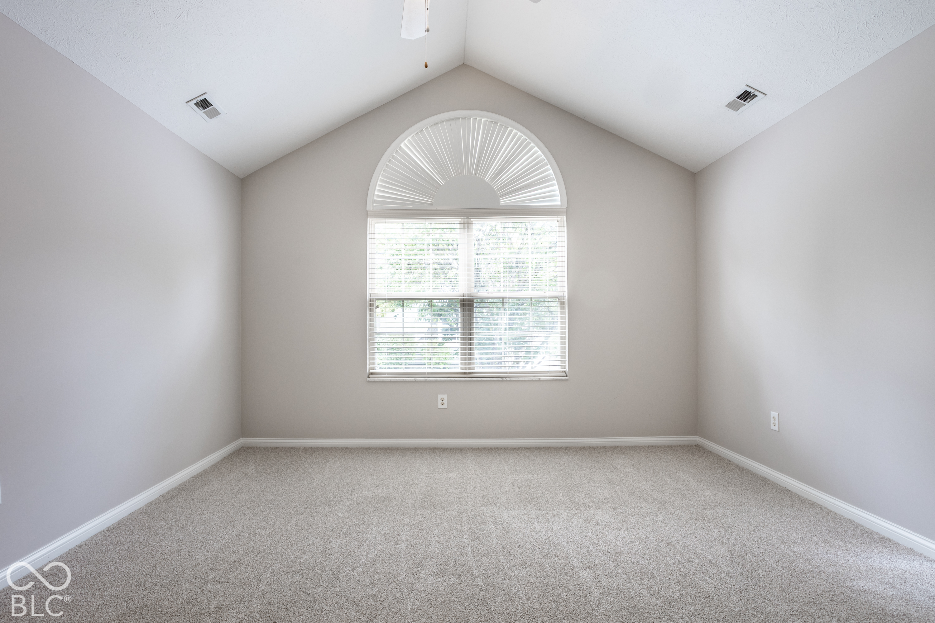 Empty carpeted room with a vaulted ceiling and a large arched window with blinds.