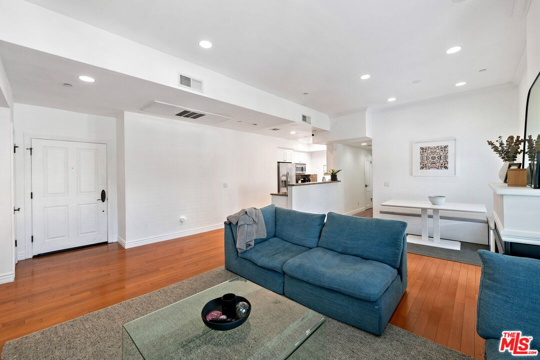 Furnished living room with a blue sofa, glass coffee table, hardwood floors, and an open kitchen.