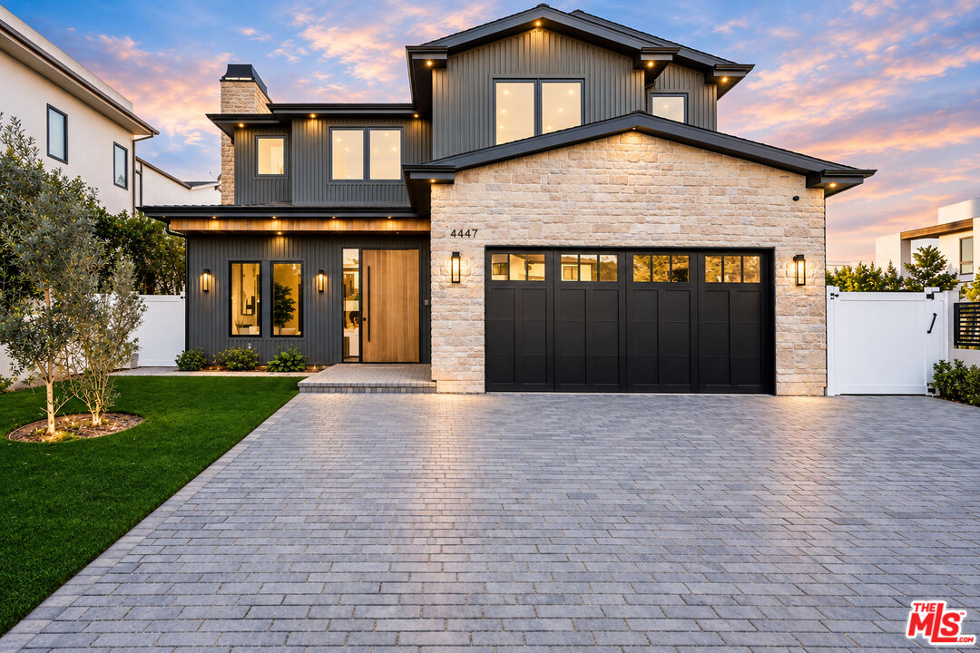 Modern two-story home with dark siding, a stone garage facade, and a wide paved driveway at sunset.