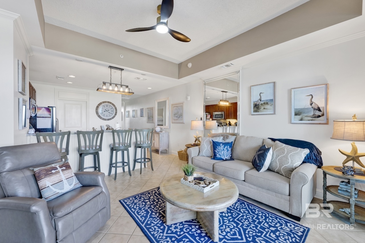 Staged living room with a gray sofa, blue patterned rug, and open dining area with teal stools.