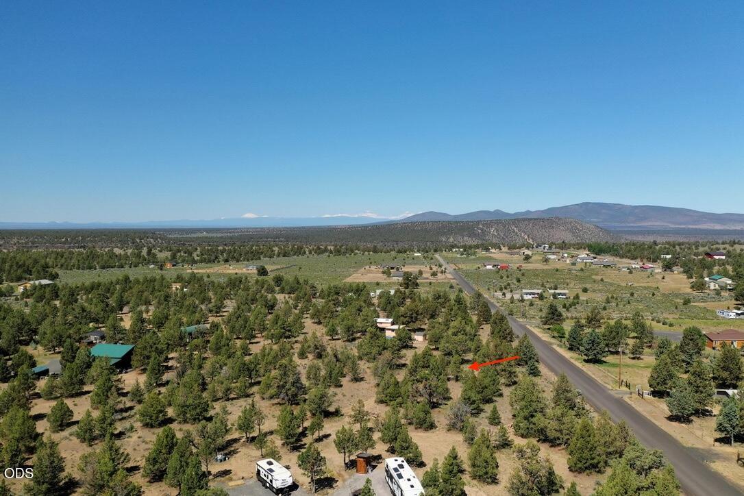 Aerial view of rural land marked by a red arrow, with juniper trees, a nearby road, and distant mountains.