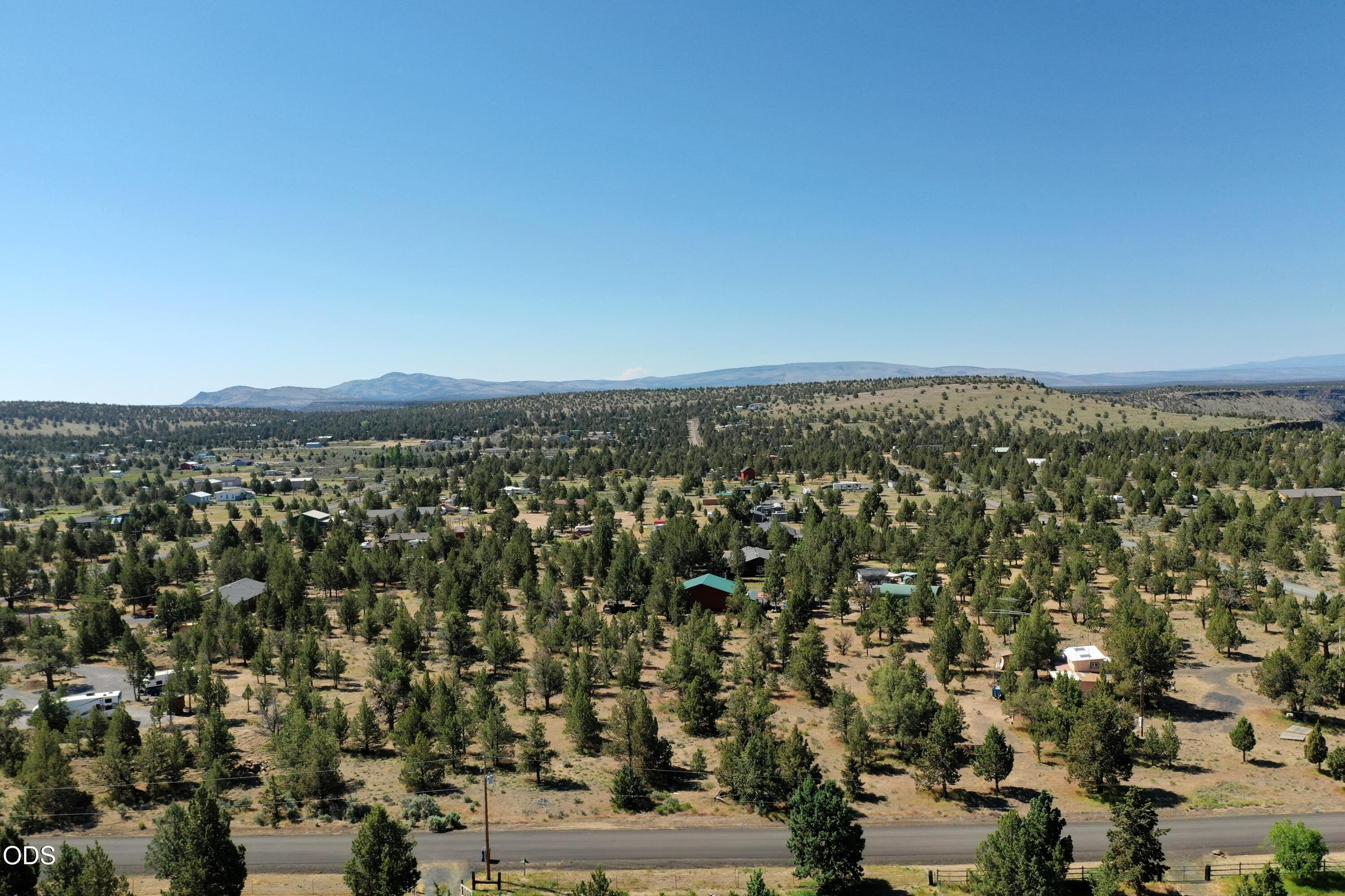 Aerial view of wooded rural land with scattered homes, a foreground road, and distant hills.