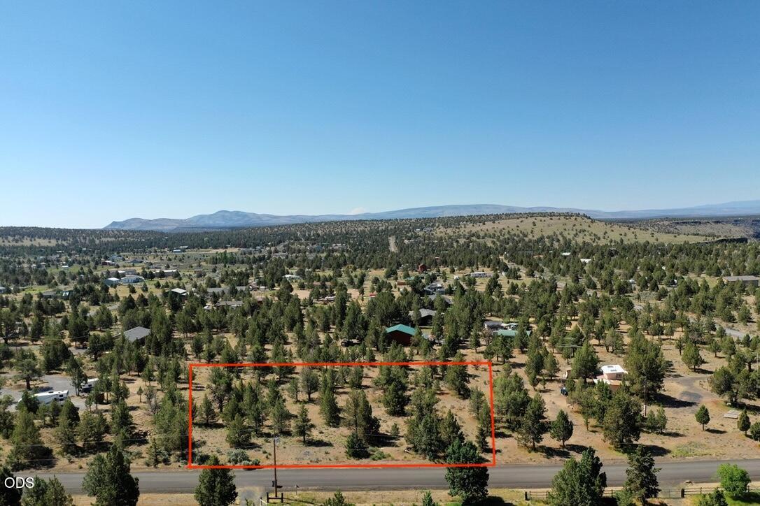 Aerial view of a vacant, tree-dotted parcel outlined in red beside a road, with distant hills.