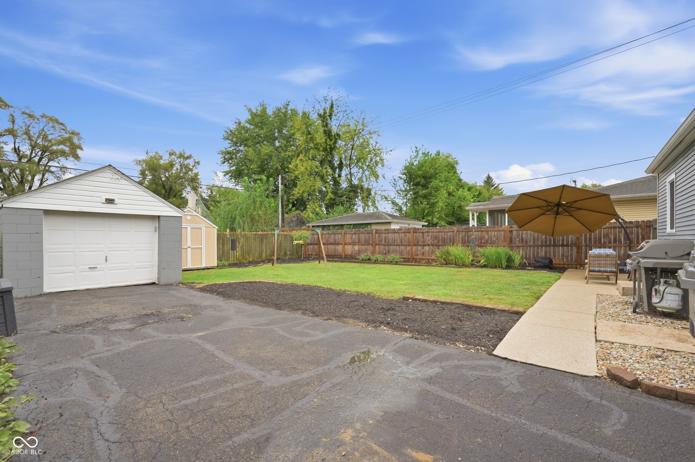 Fenced backyard with a lawn, detached garage, and patio umbrella beside a paved driveway.
