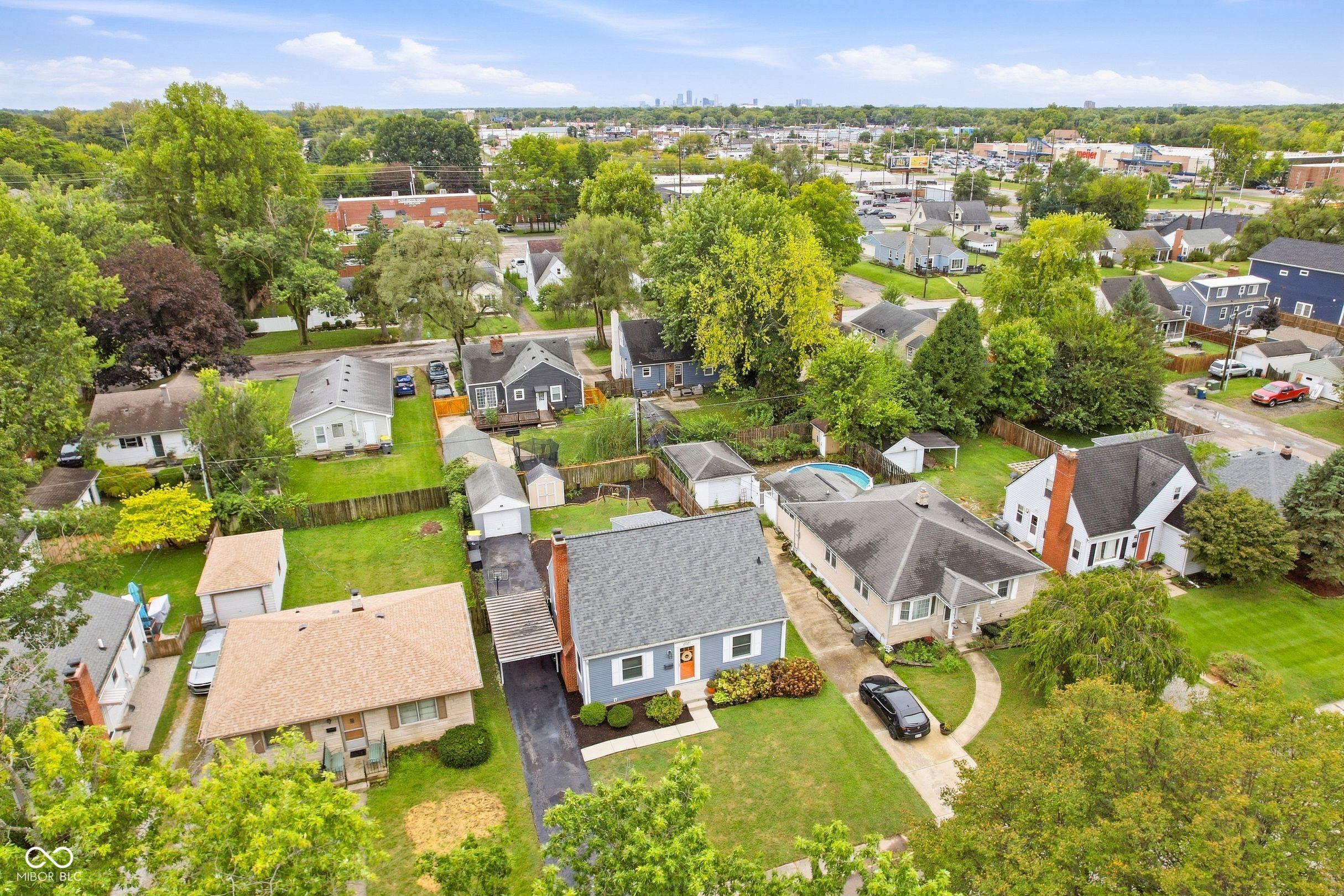 Aerial view of a blue single-story house with a front garden, driveway, and Indianapolis skyline beyond the neighborhood.