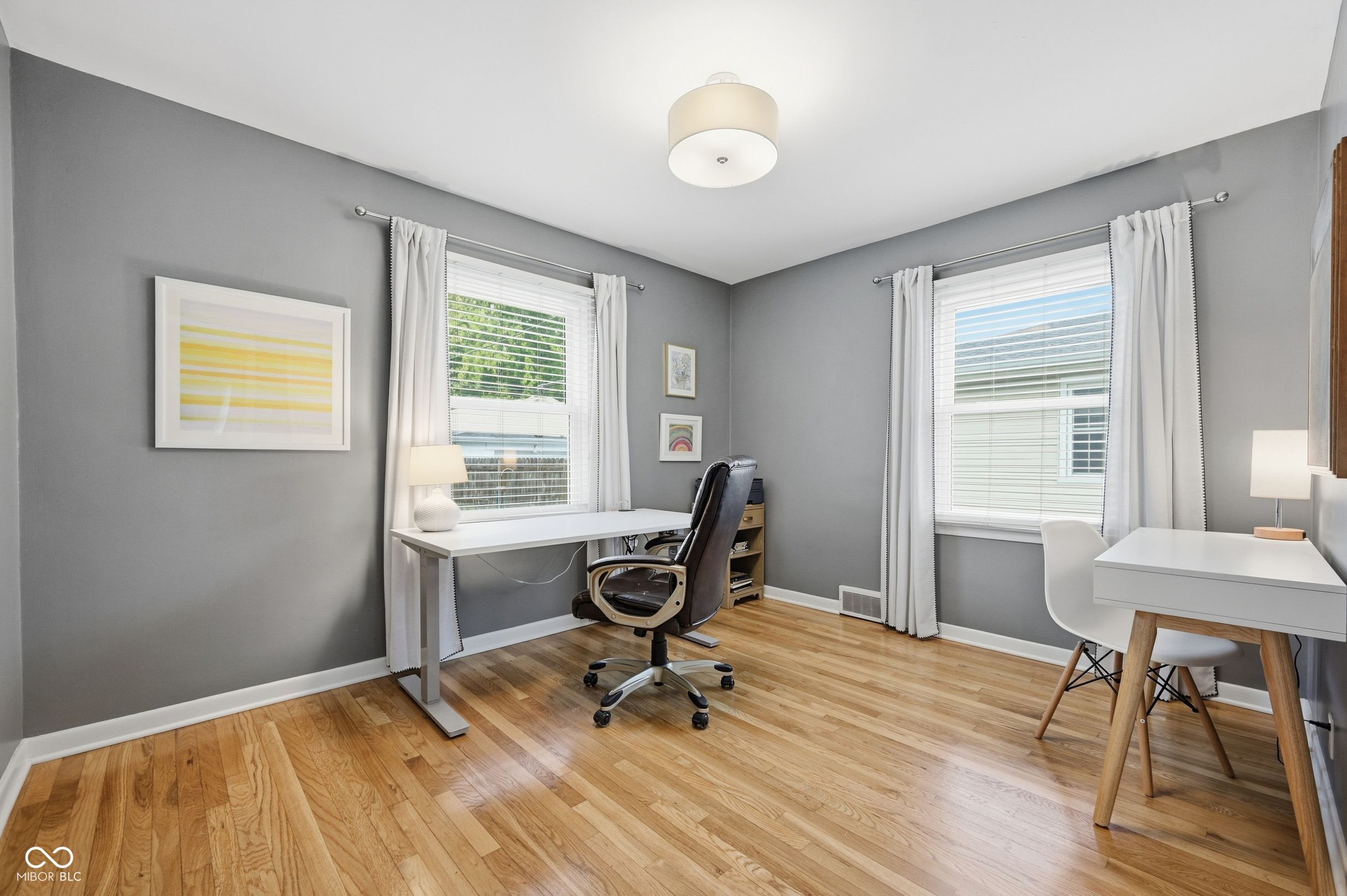 Home office with gray walls, hardwood floors, two windows, and white desks.