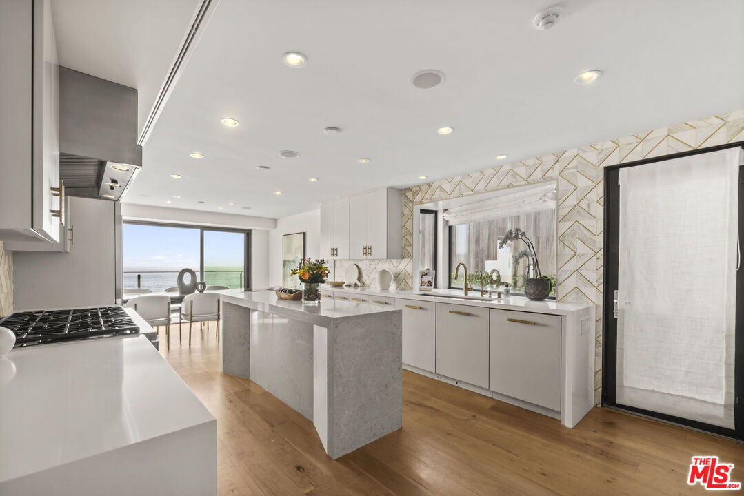 Modern white kitchen with a large stone island and ocean views through sliding glass doors.