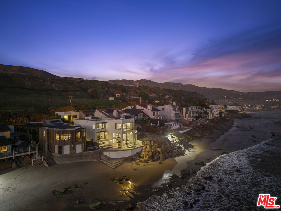 Modern beachfront homes glow at dusk along Malibu’s rocky, wave-washed shoreline.