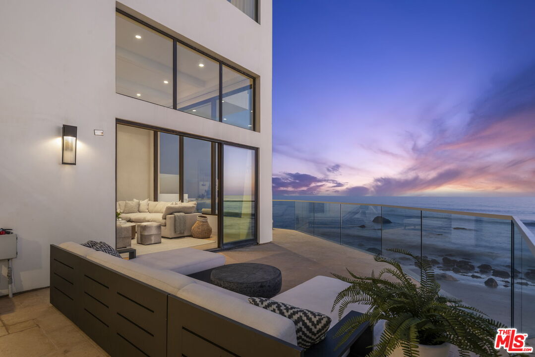 Modern oceanfront home terrace with lounge seating and glass railing overlooking the Pacific at dusk.