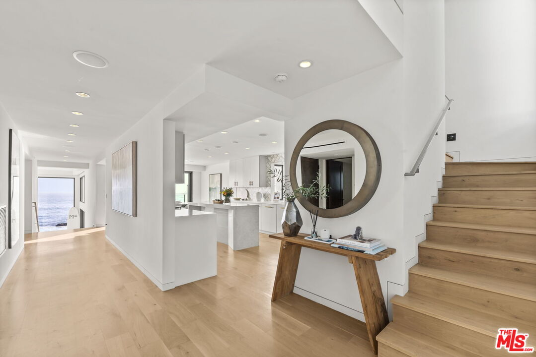 Open entry hall with pale wood floors, a round mirror, wooden staircase, and ocean view down the hall.