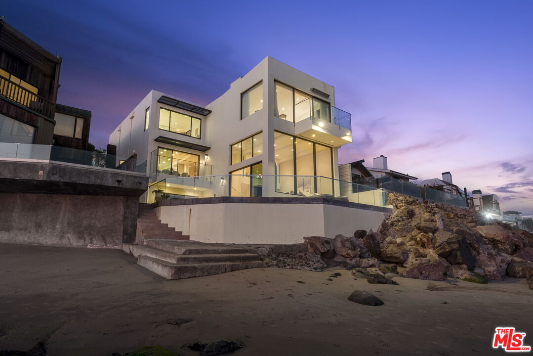 Modern beachfront home with glass balconies above a rocky, sandy shore at dusk.