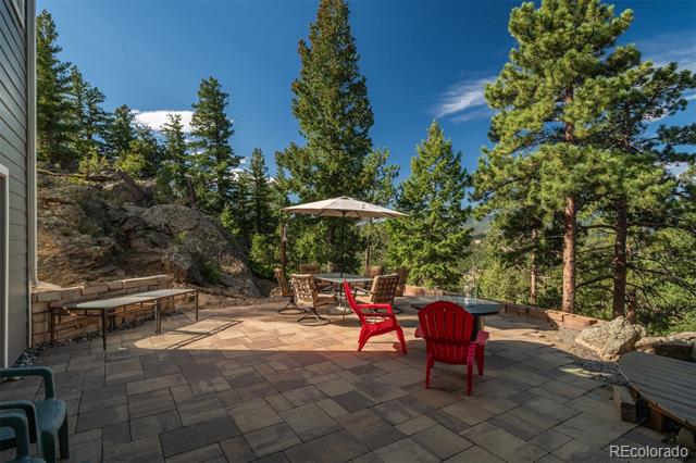 Stone patio with red Adirondack chairs and dining set among pine trees.