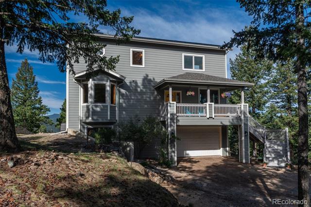 Gray two-story house with a covered porch, elevated garage, and stairs among pine trees.