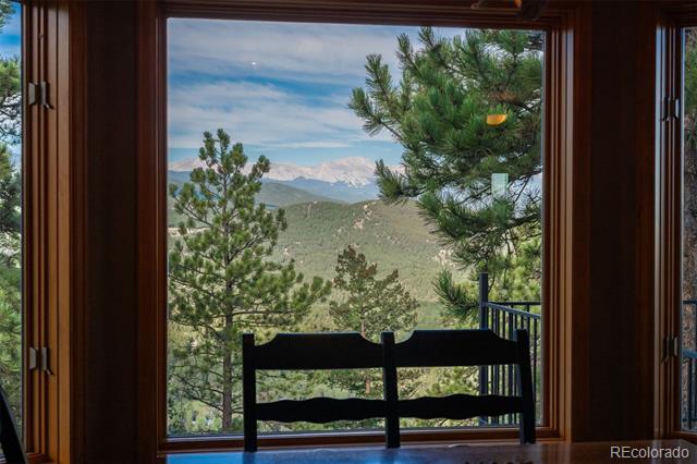 Mountain view through a large wood-framed window, with pine trees and a chair in the foreground.
