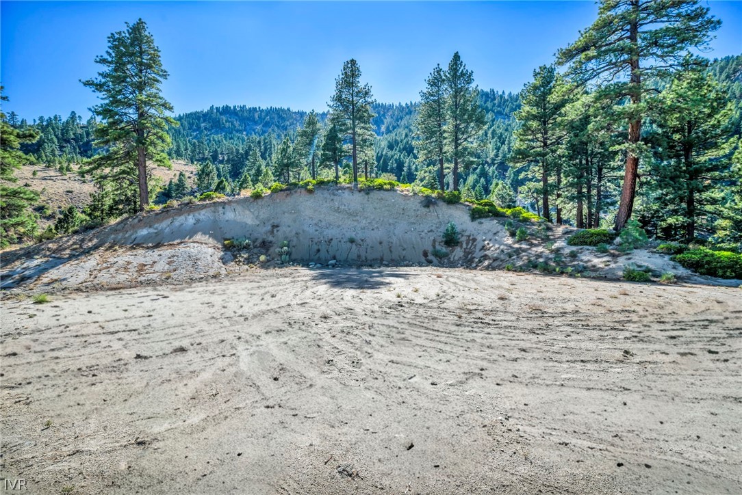 Vacant mountain lot with a broad graded dirt clearing, pine trees, and wooded hills.