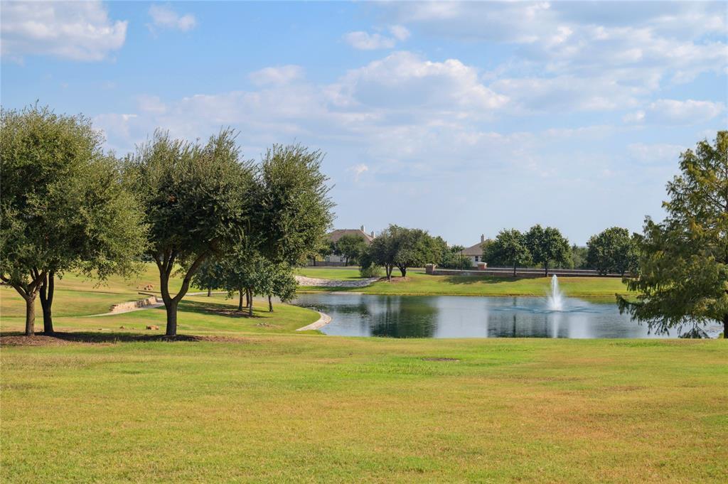 Landscaped park pond with a fountain, curving shoreline, mature trees, and broad grassy lawn.