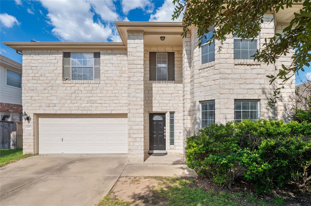 Two-story stone house with an attached garage, dark front door, and leafy landscaping.