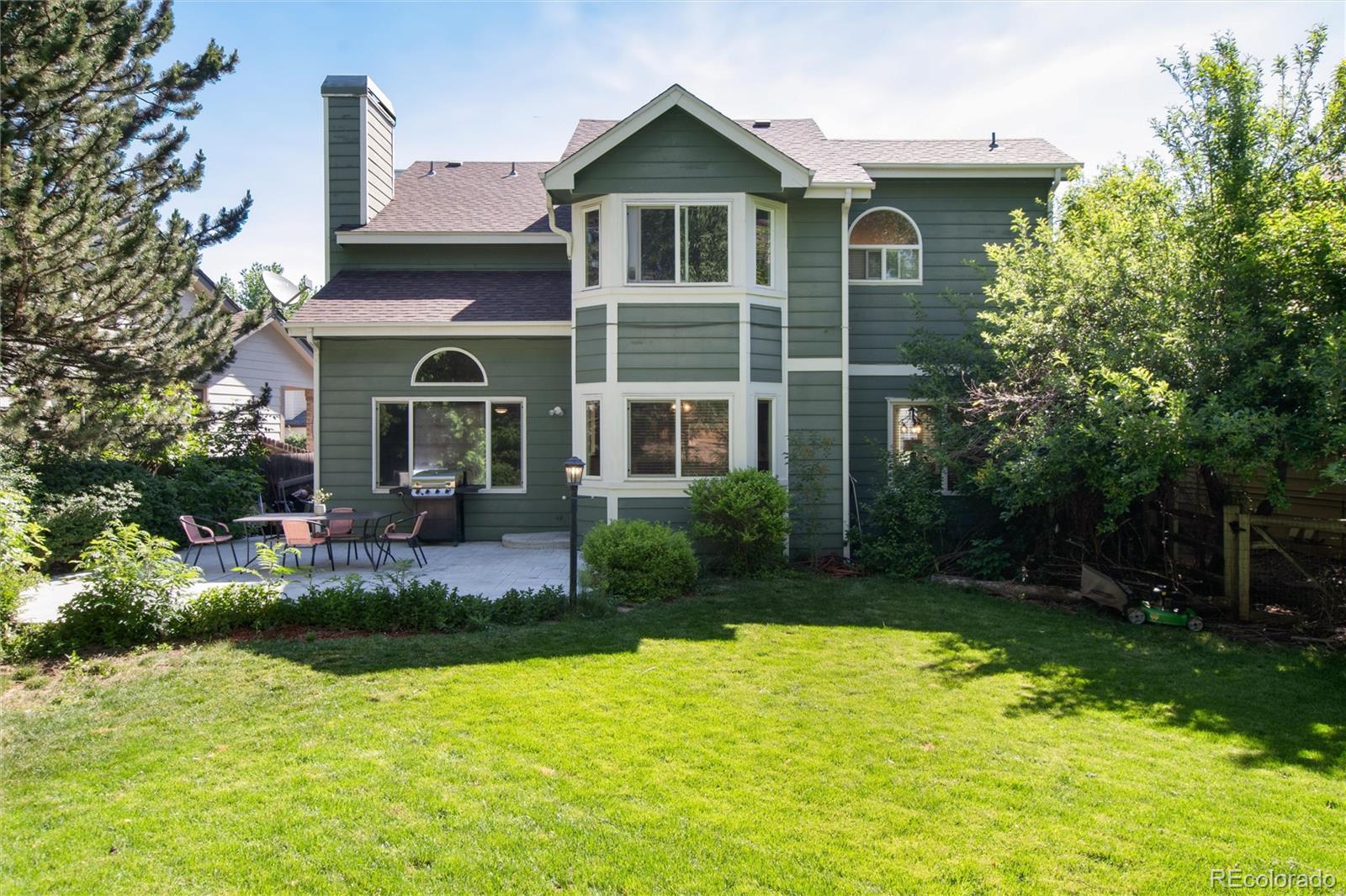 Green two-story home with a paved patio, outdoor dining set, grill, and broad sunny lawn.