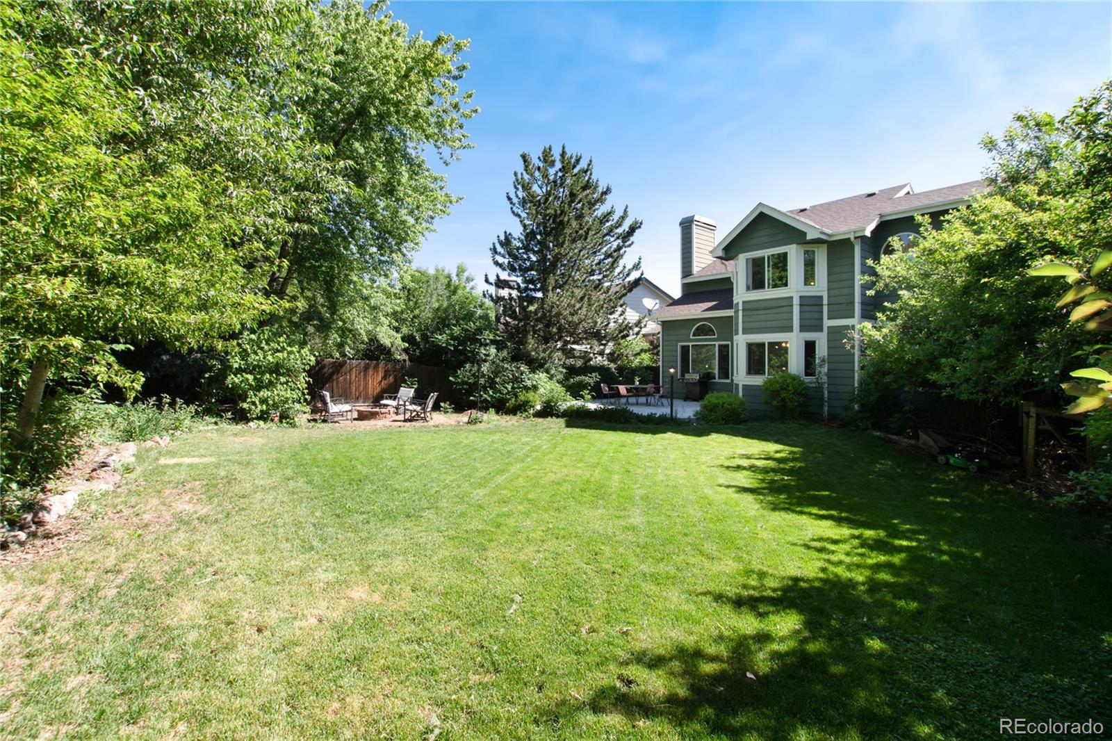 Two-story green house beside a spacious, tree-lined lawn and patio seating.