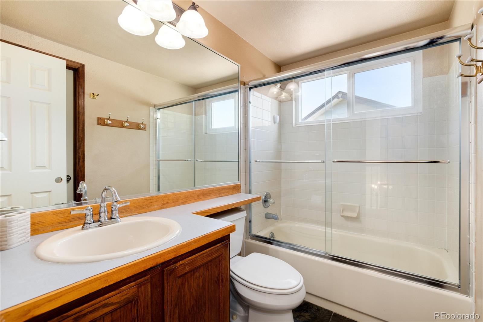 Bathroom with a wood vanity, large mirror, and glass-enclosed bathtub beneath a wide window.