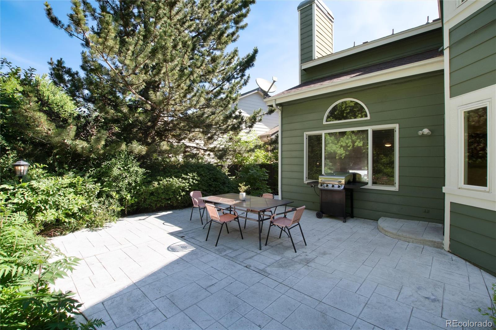 Stone-paver patio beside a green house, furnished with outdoor dining set and grill.