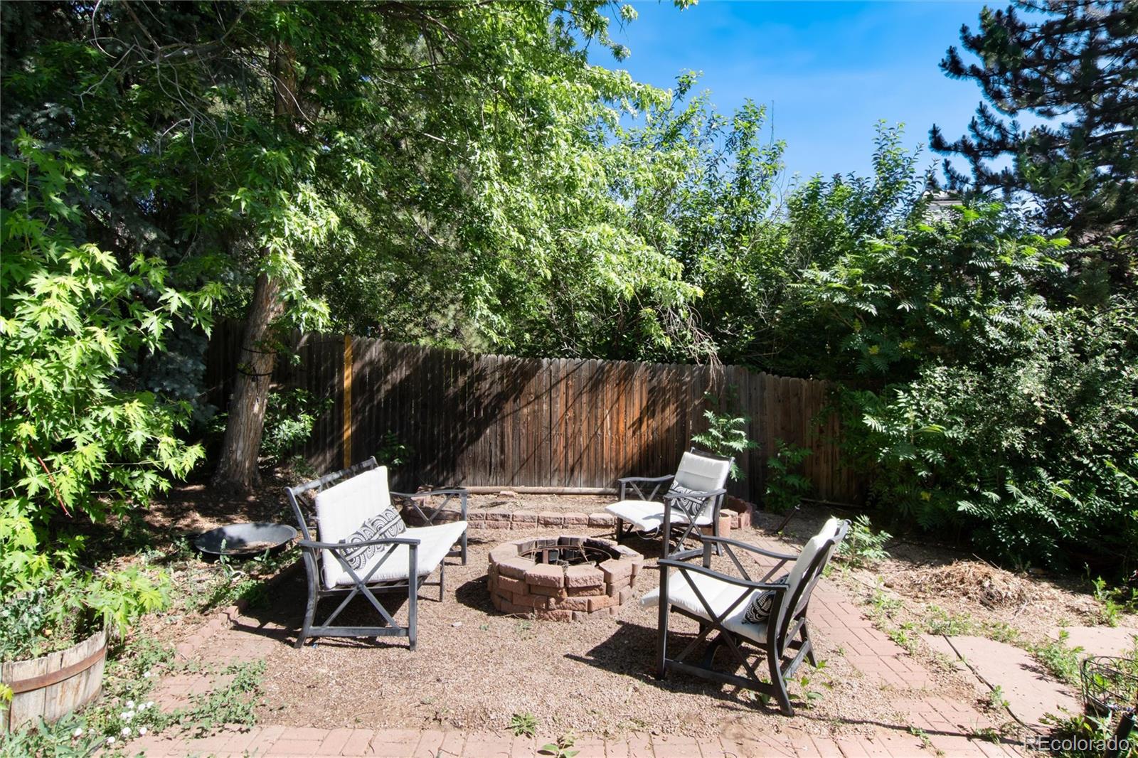 Backyard patio with a stone fire pit, cushioned chairs, brick paving, and mature trees.
