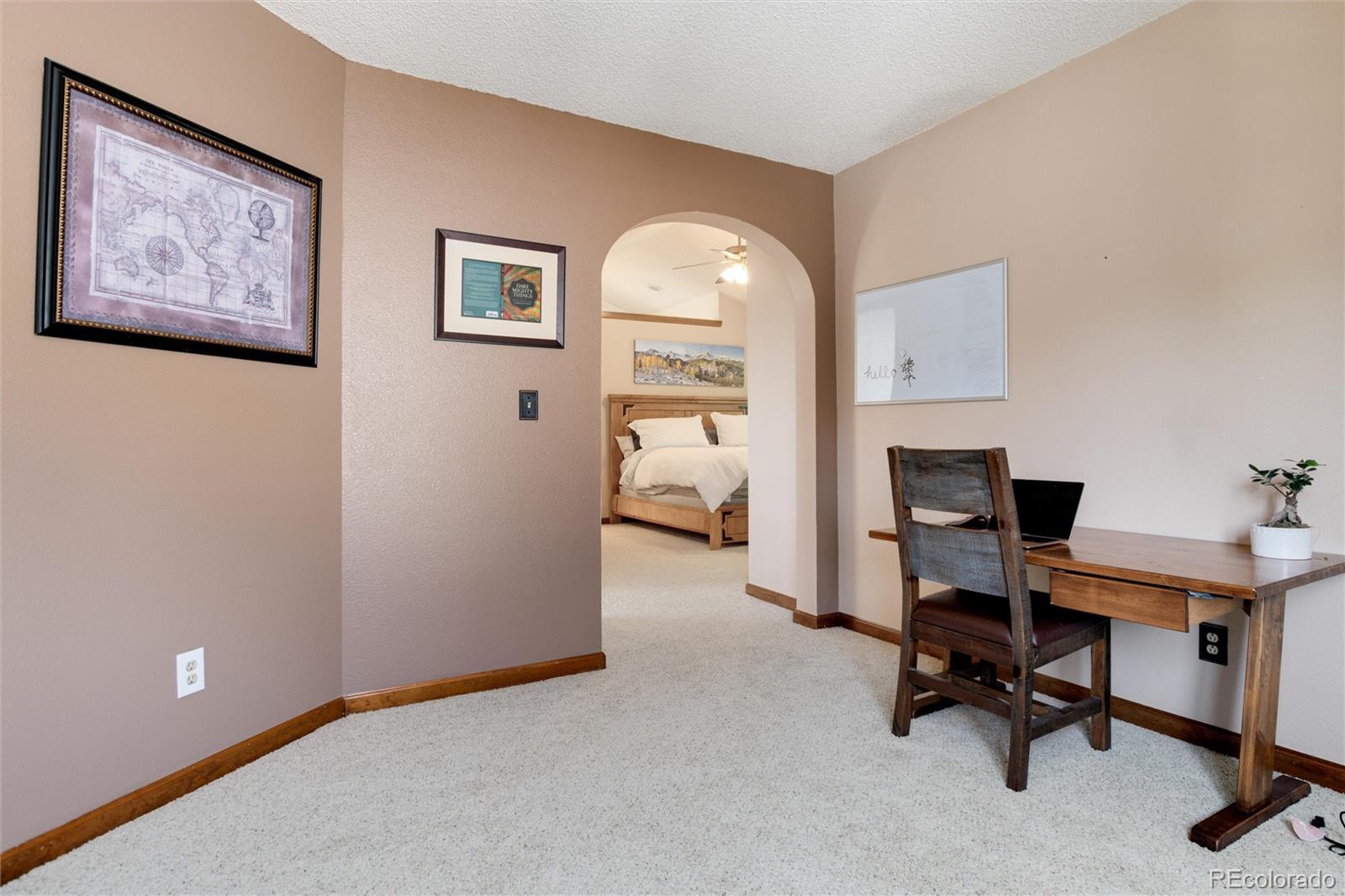 Carpeted room with a wooden desk beside an archway opening into a bedroom.