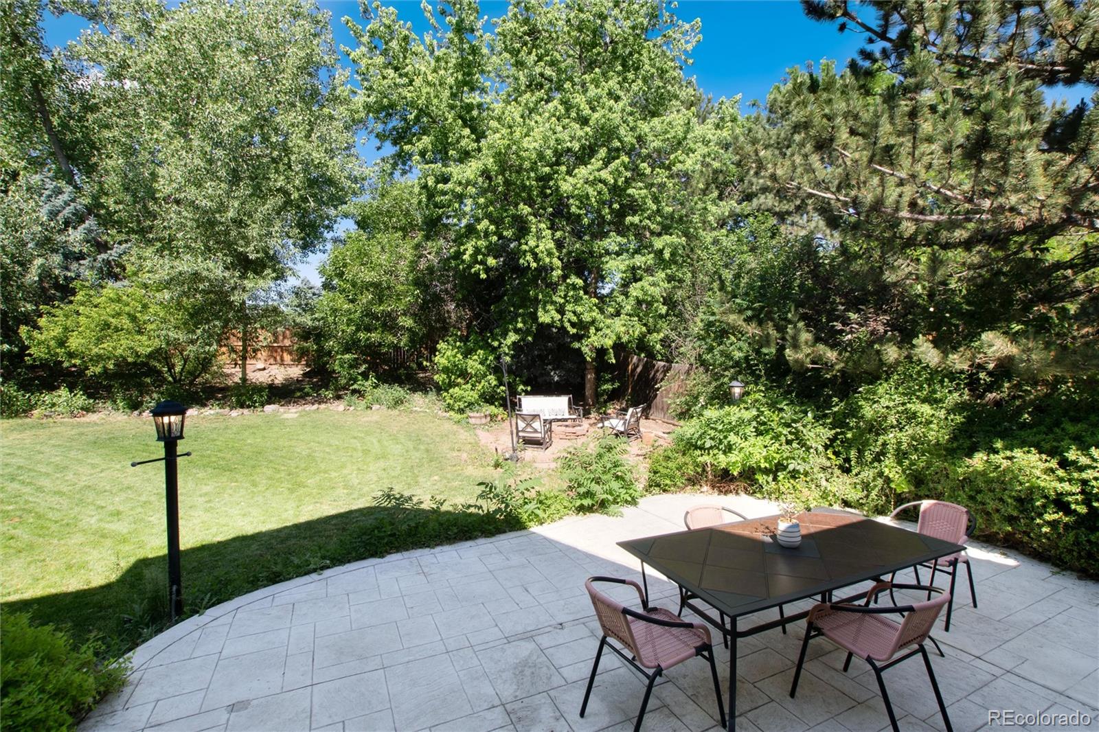 Paved backyard patio with outdoor dining set beside a lawn, mature trees, and garden seating.