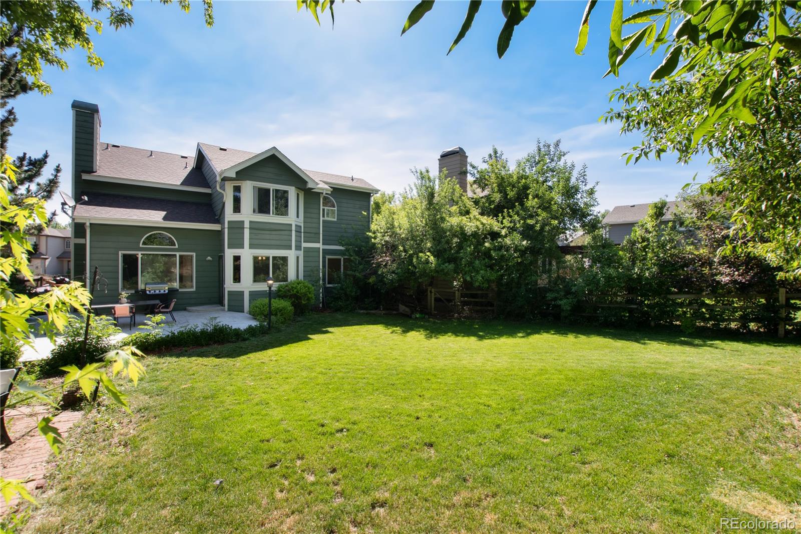 Green two-story home with a patio and grill opening onto a broad, tree-bordered lawn.