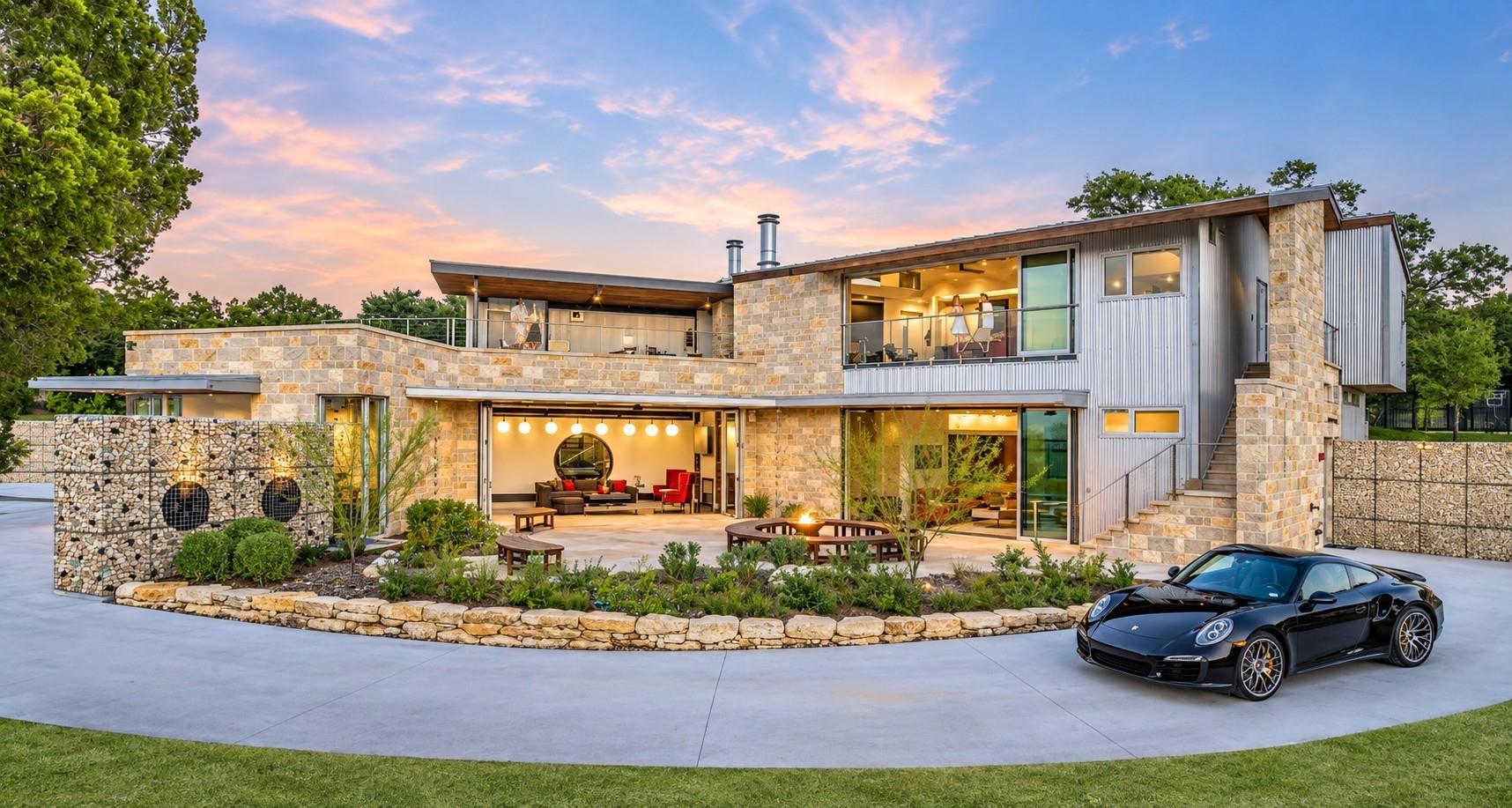 Modern stone-and-metal home with illuminated open living areas and a black sports car in the driveway.