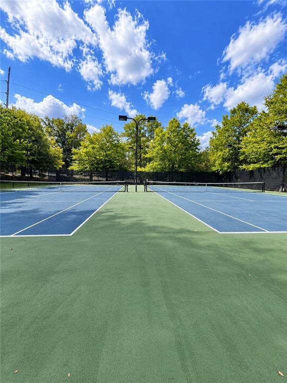 Outdoor tennis court with blue playing areas, green surface, surrounding trees, and a bright, cloud-dotted sky.