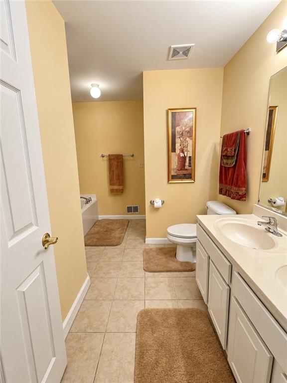 Bathroom with a double-sink vanity, toilet, beige tile floor, and brown bath mats.