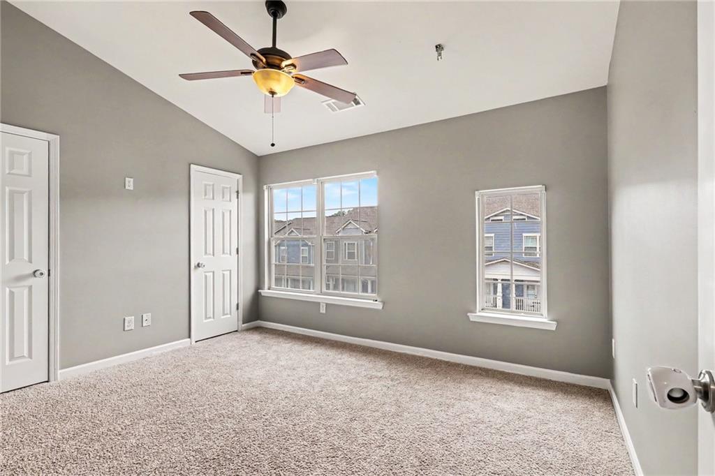 Empty bedroom with gray walls, carpet, ceiling fan, and windows facing neighboring homes.