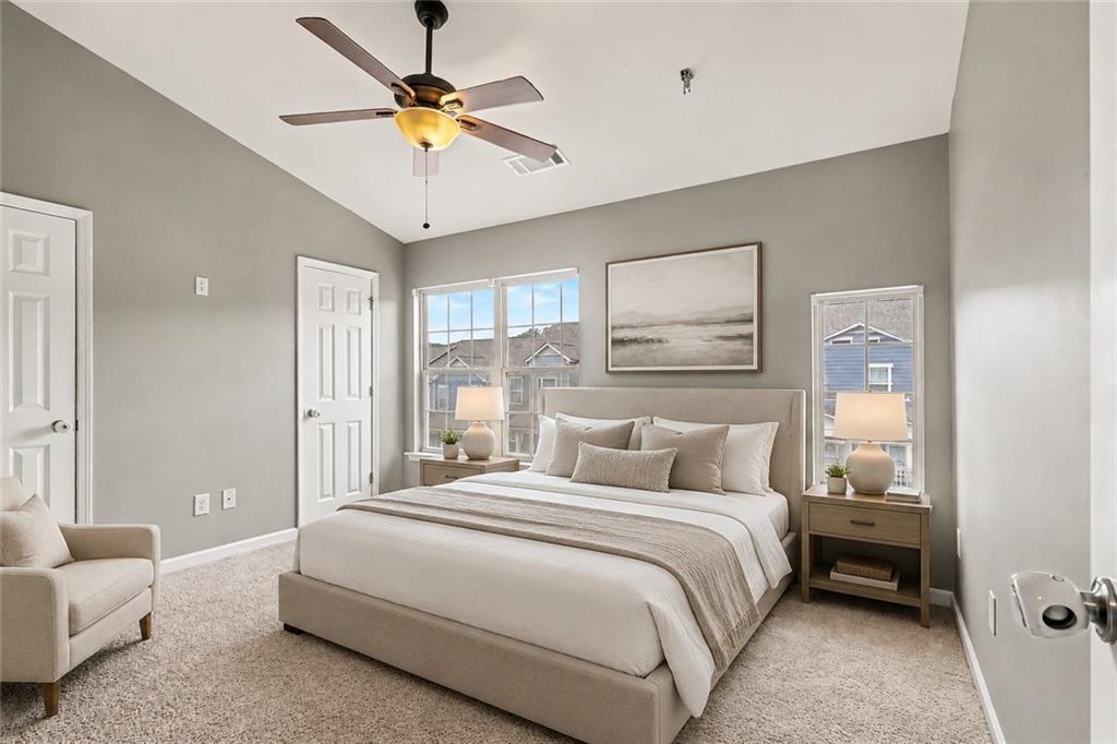 Staged bedroom with a king bed, ceiling fan, and windows overlooking neighboring homes.