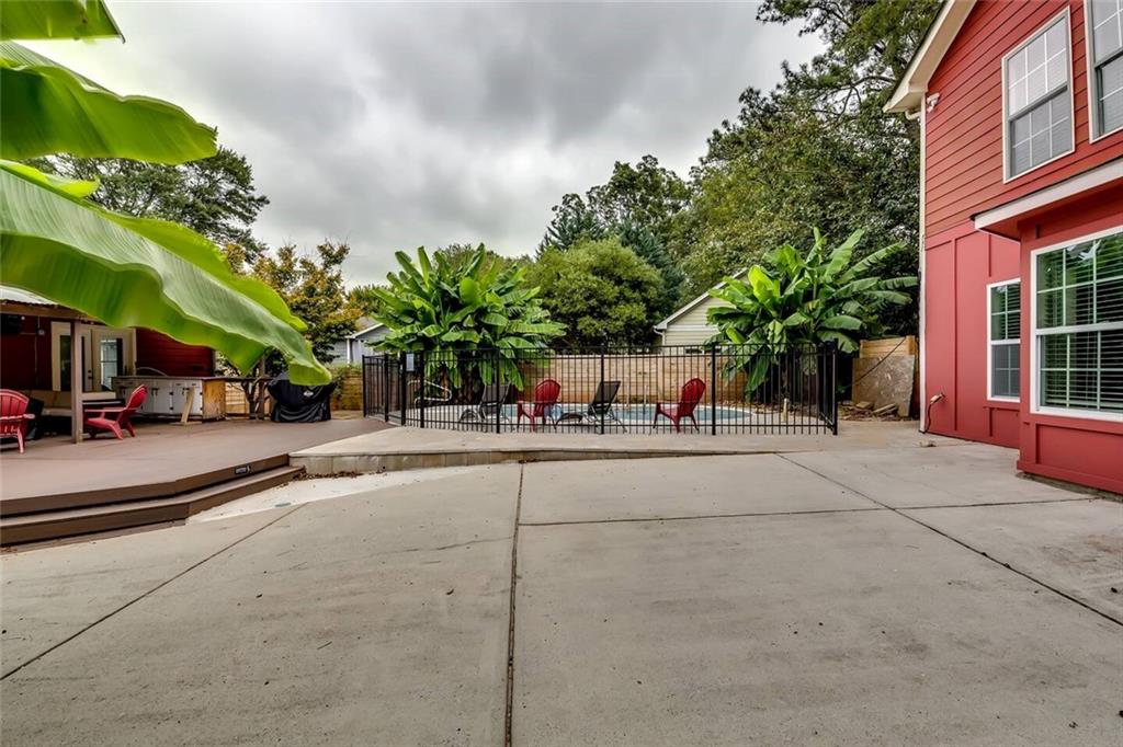 Backyard patio with a fenced seating area, concrete courtyard, and leafy banana plants beside a red-sided house.