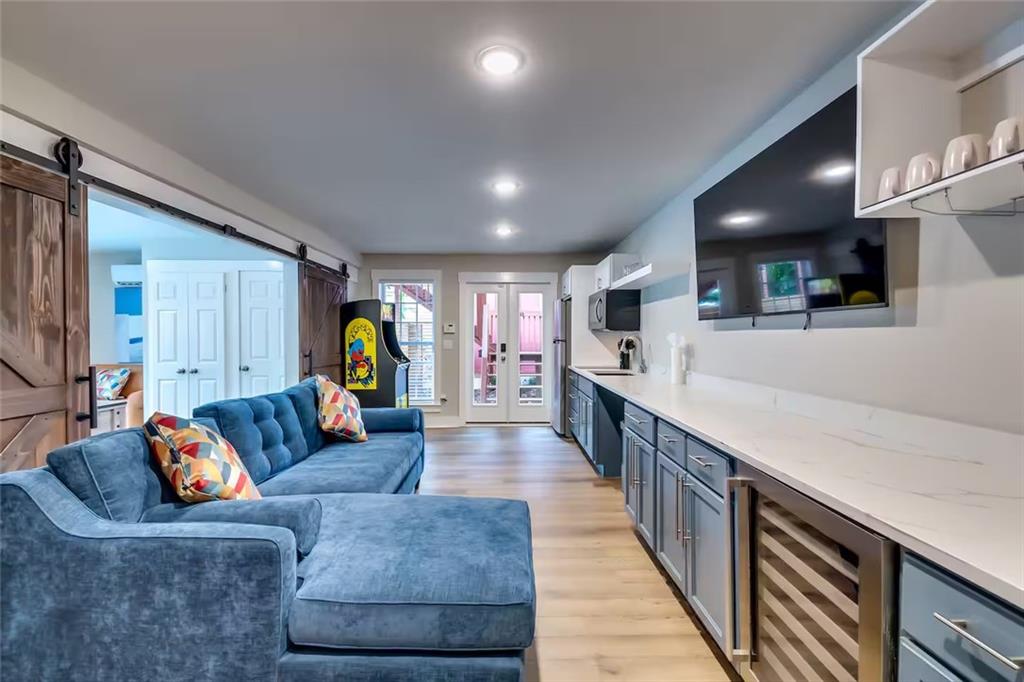 Furnished living room with a blue sectional, long kitchenette counter, wall-mounted TV, and arcade machine.