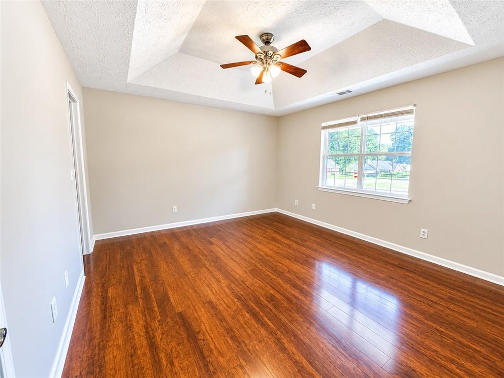 Empty room with warm wood flooring, a tray ceiling, ceiling fan, and large window.